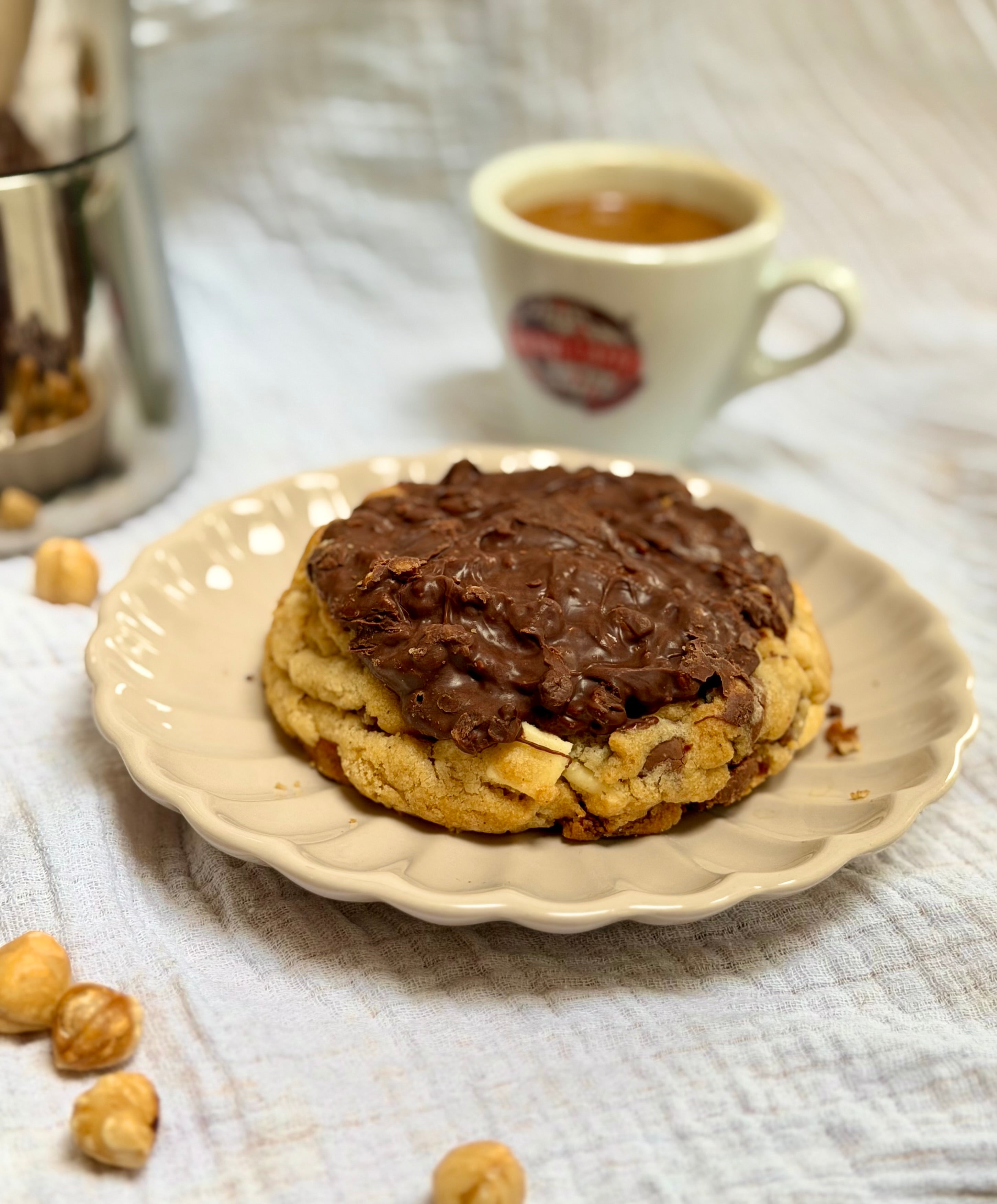 A chocolate-topped cookie on a scalloped cream plate, with hazelnuts scattered around and a white coffee cup in the soft-focus background on a textured white cloth.