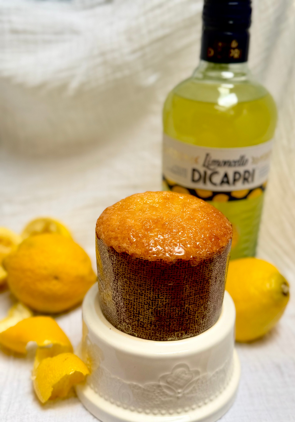 A limoncello cake on a white pedestal dish, with a bottle of Limoncello DiCapri lemon liqueur and fresh lemons in the background on a white textured fabric.