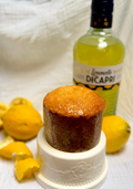 A limoncello cake on a white pedestal dish, with a bottle of Limoncello DiCapri lemon liqueur and fresh lemons in the background on a white textured fabric.