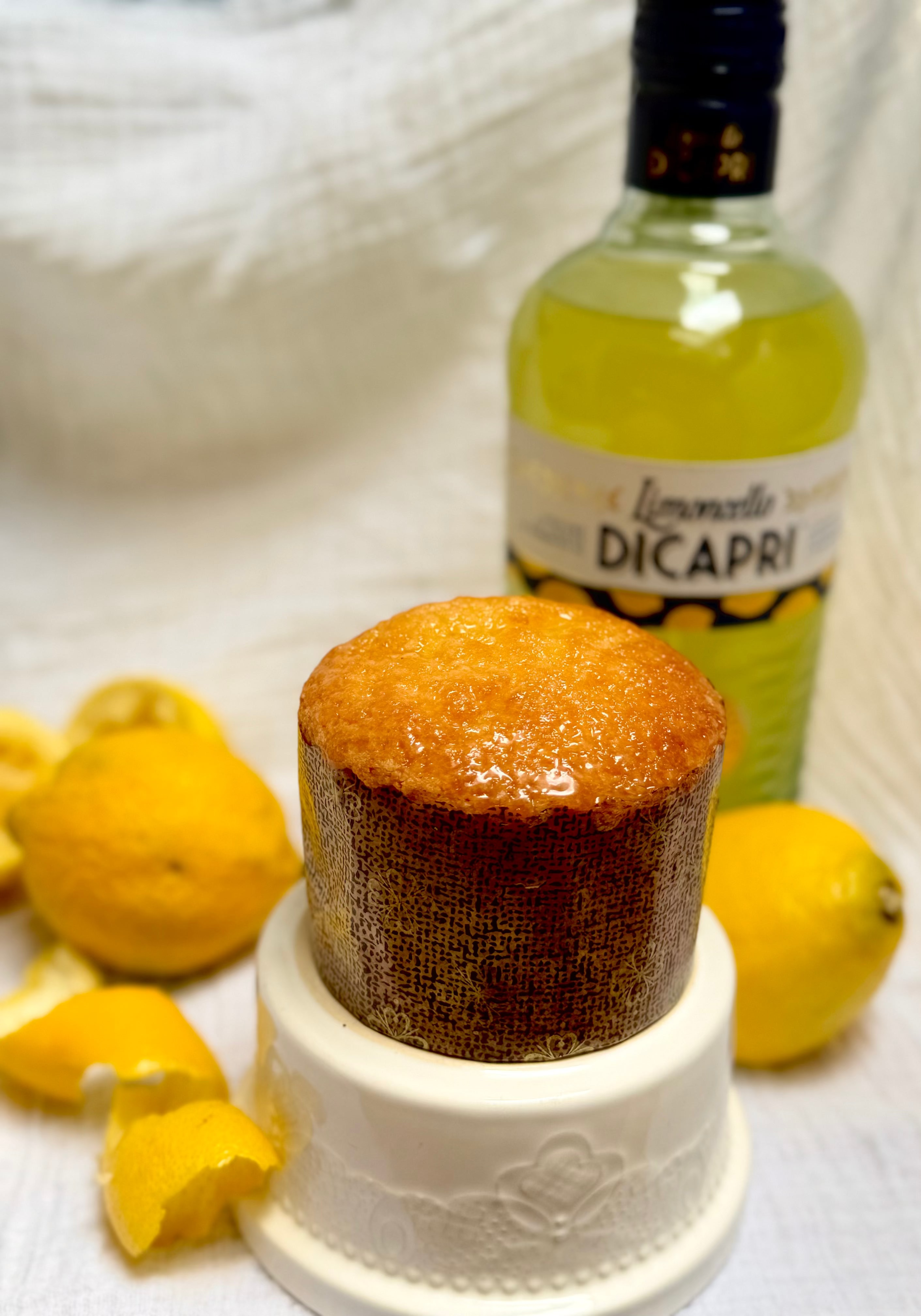 A limoncello cake on a white pedestal dish, with a bottle of Limoncello DiCapri lemon liqueur and fresh lemons in the background on a white textured fabric.